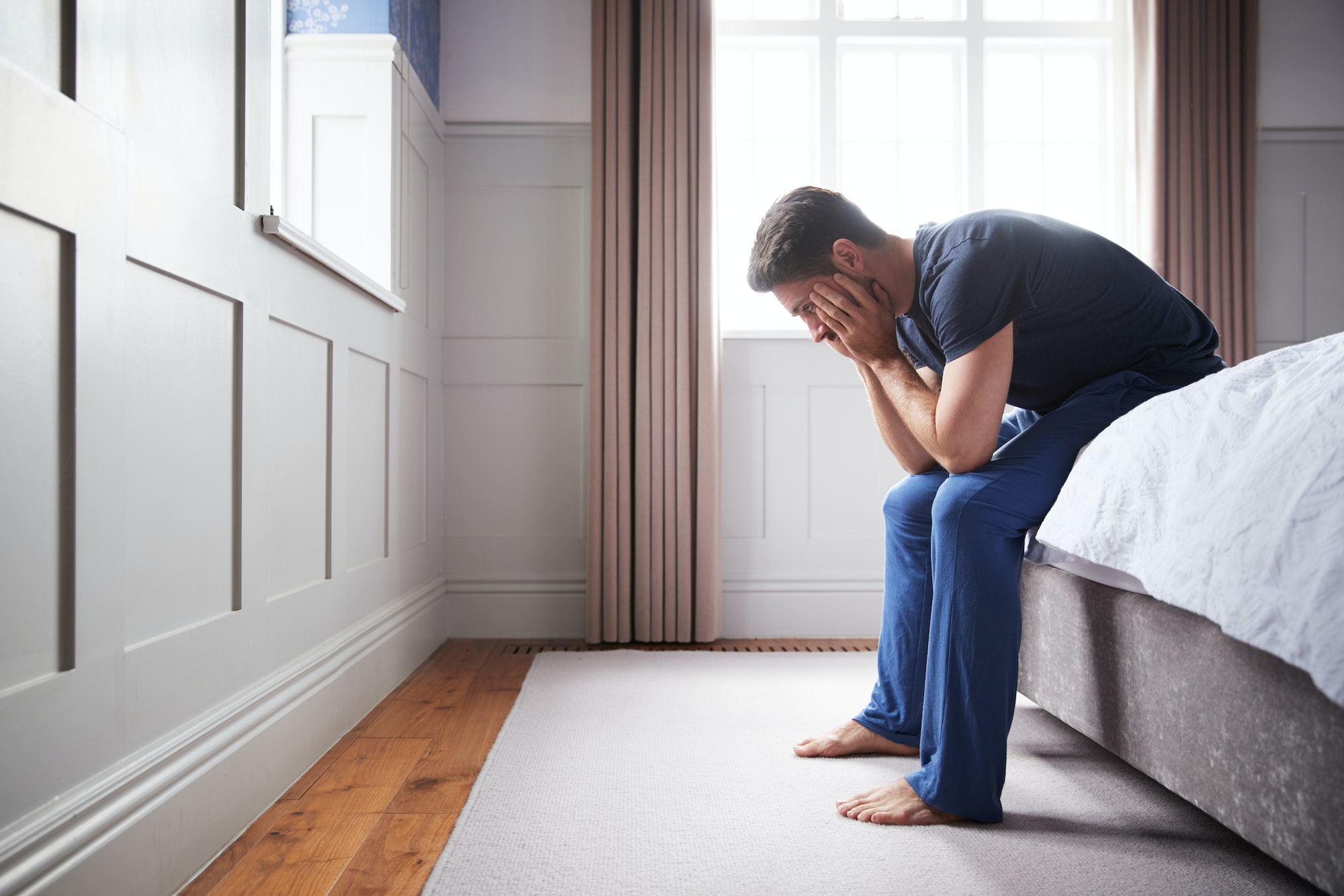 Man Wearing Pajamas Suffering With Depression Sitting On Bed At Home